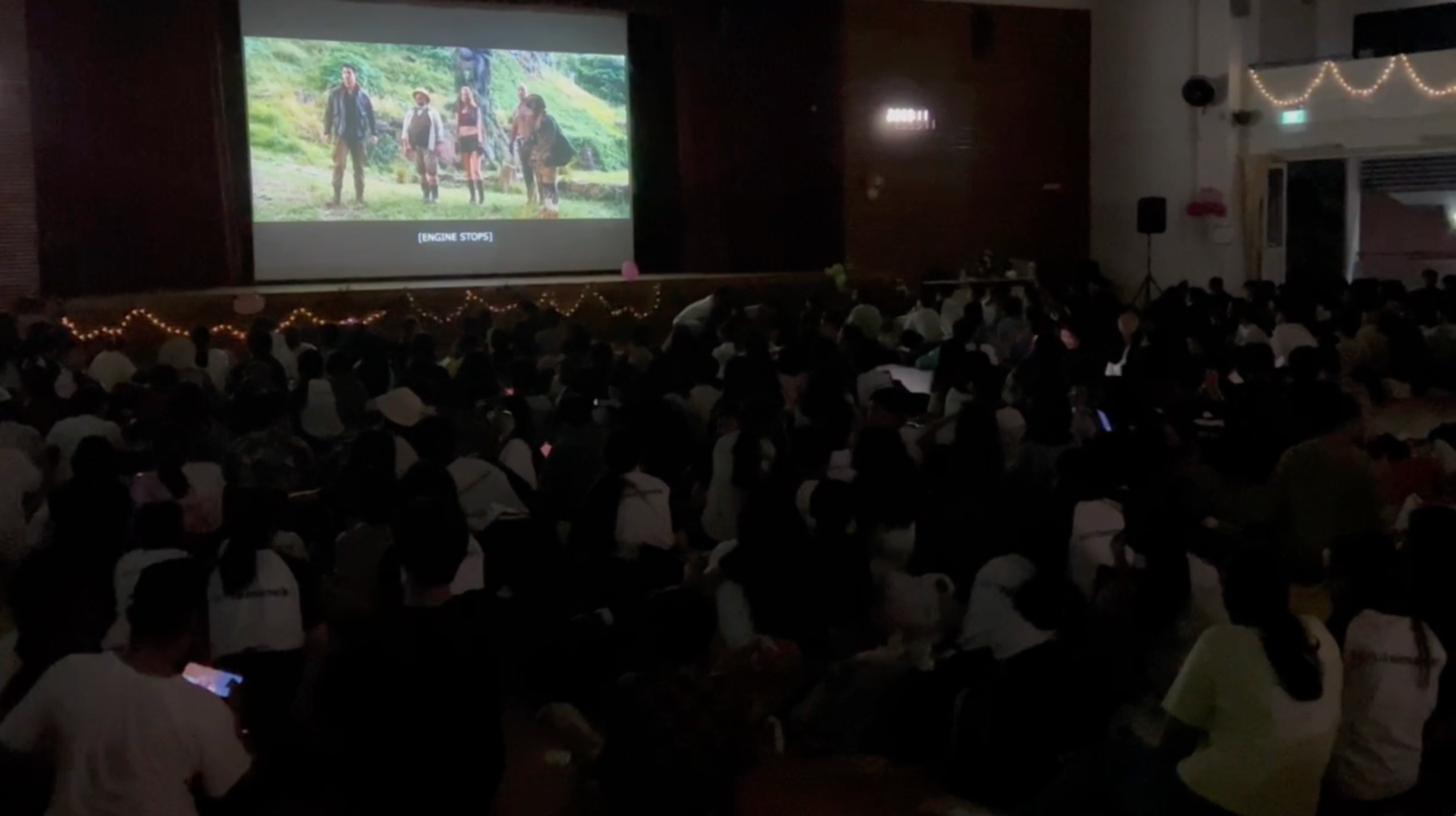 Large crowd seated on a floor in a dark hall watching a movie projected on a screen at the front.